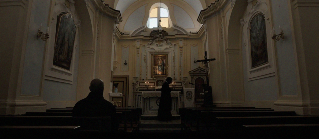 McCall sentado no banco da igreja, em frente ao altar, onde encontra-se um padre.